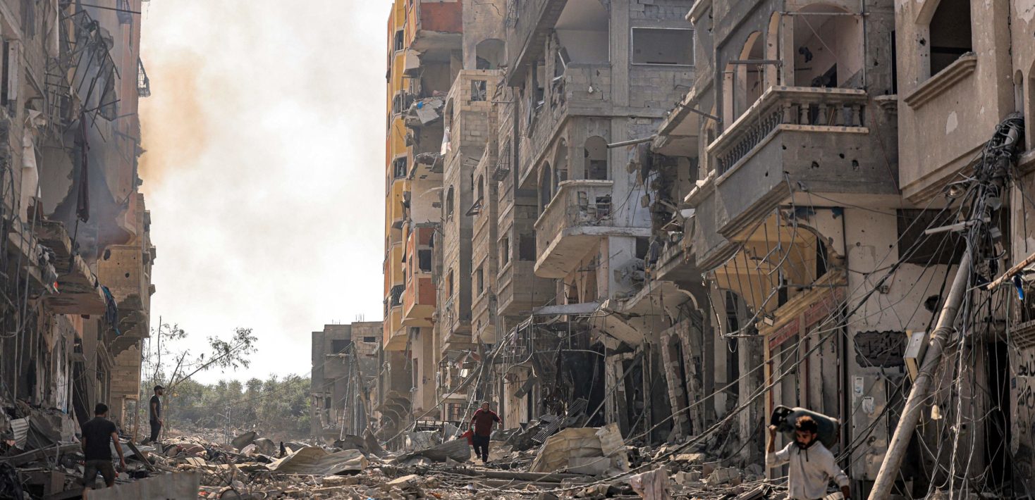 Photo of a street littered with debris and rubble. On both sides, buildings are heavily damaged. A man walks through the street carrying a propane tank.