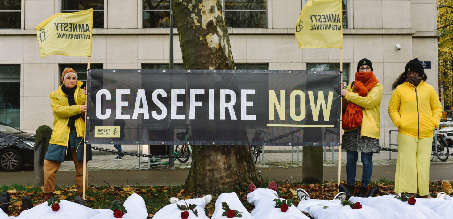 Photo taken in front of the USA embassy in Brussels. Three persons are standing holding a large banner, reading "Ceasefire now", along with two yellow flags which read "Amnesty International" with the candle logo. Below them, 9 persons are lying on the ground wrapped in white sheets, with a rose on top of each person.
