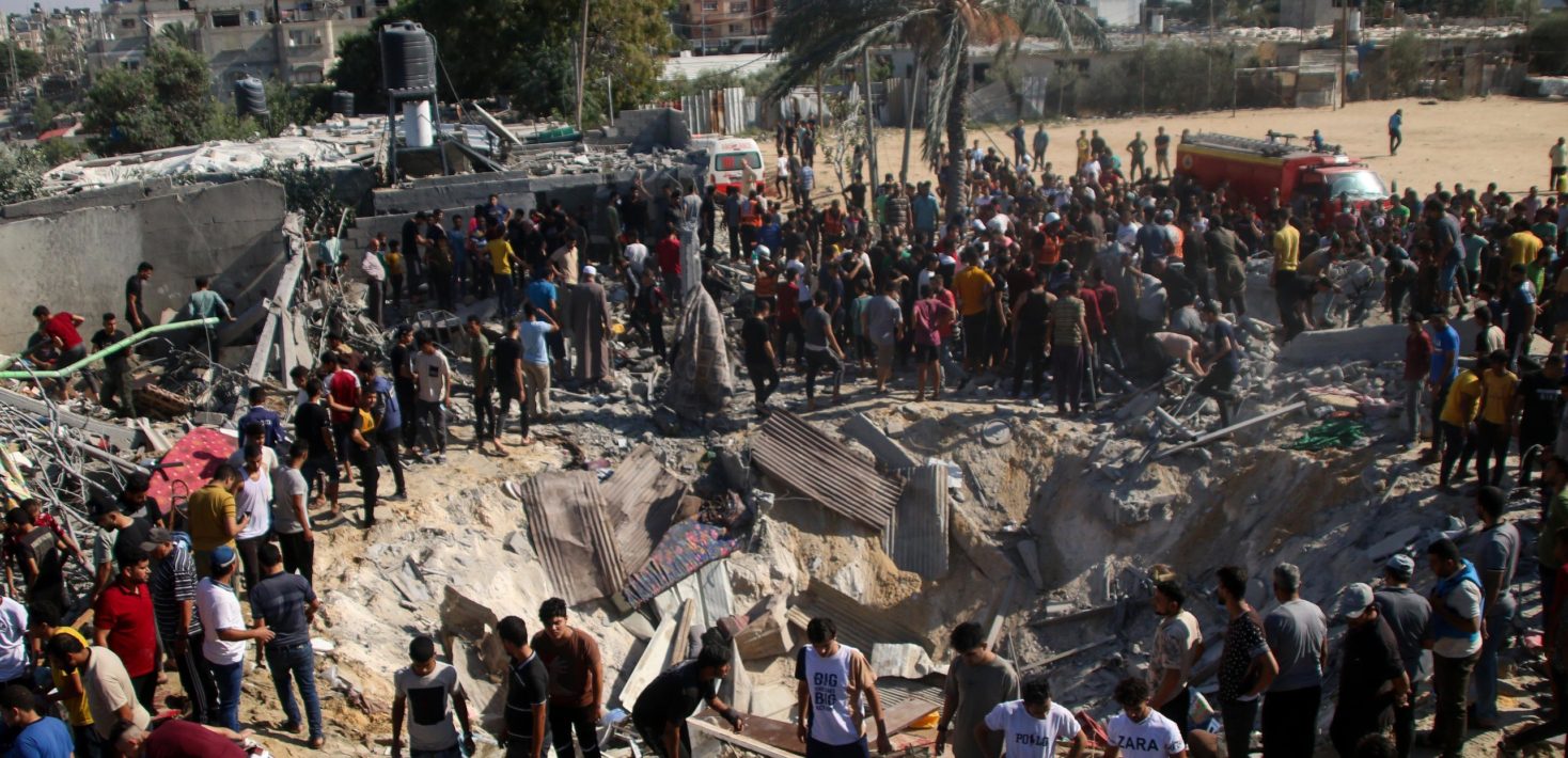 Dozens of persons surround a crater with rubble. Emergency vehicles are visible in the background of the photo.