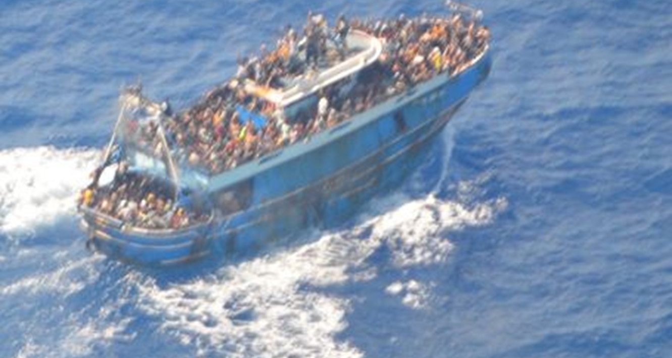 An overcrowded blue boat is visible in a blue sea. The image is an aerial view. White foam behind it indicates that it is moving. People are visible on every deck.