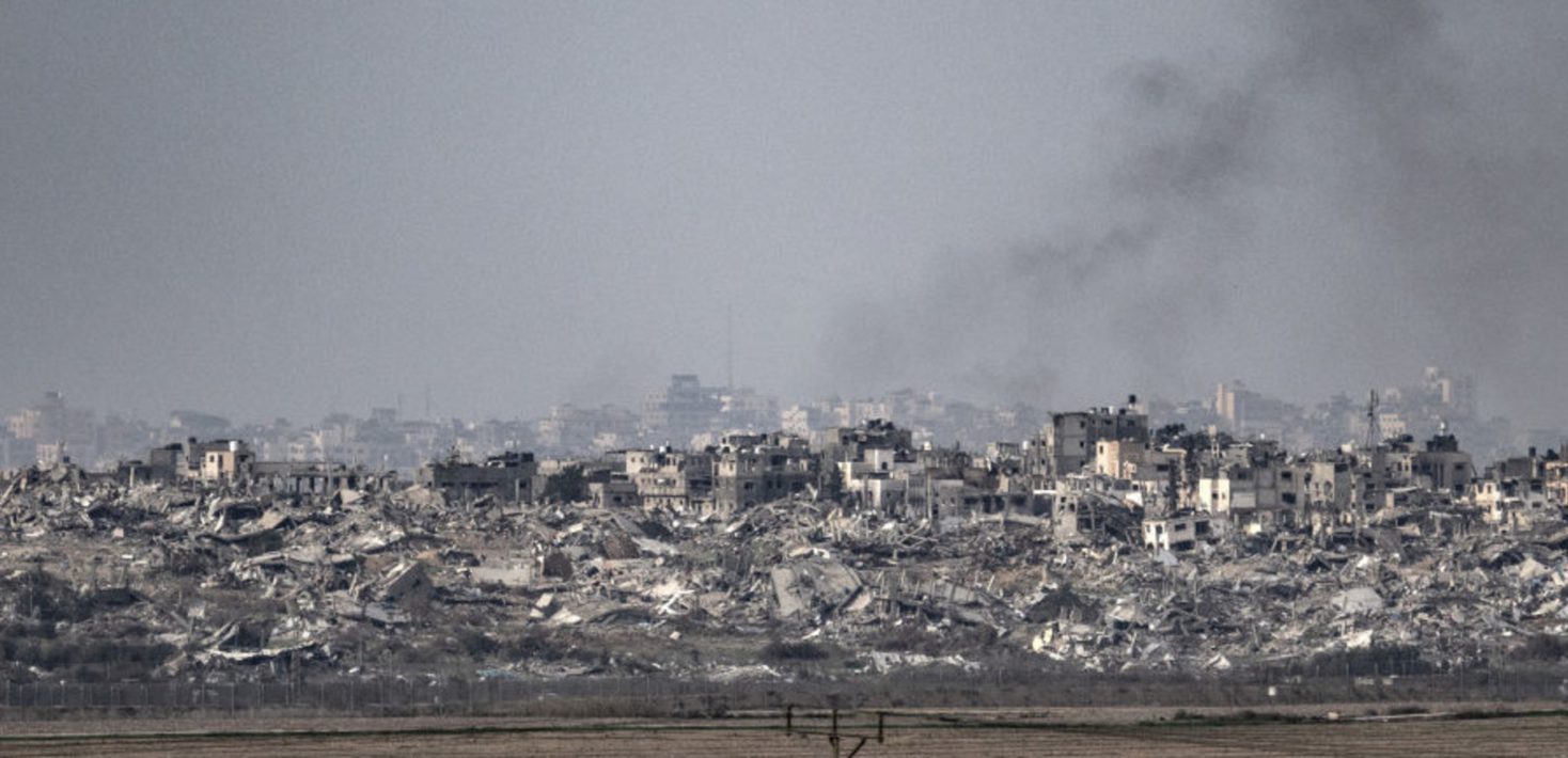 In the forefront, a stretch of empty land is visible, until a fence. Behind the fence, many destroyed buildings are visible, with smoke rising from those in the background.