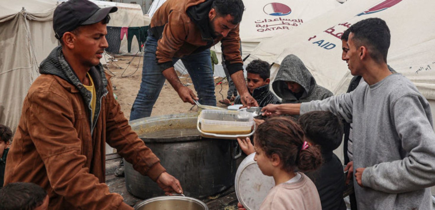 Two men are distributing soup to a group of children and men. Behind them, white tents are visible, reading "aid".